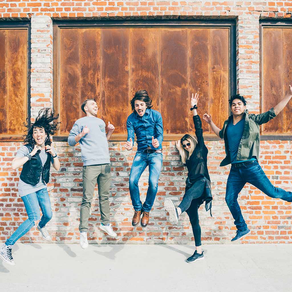 Five people jumping happily in the air. Some of them have thrown their arms up and are smiling. The background is brick wall with boarded up windows.