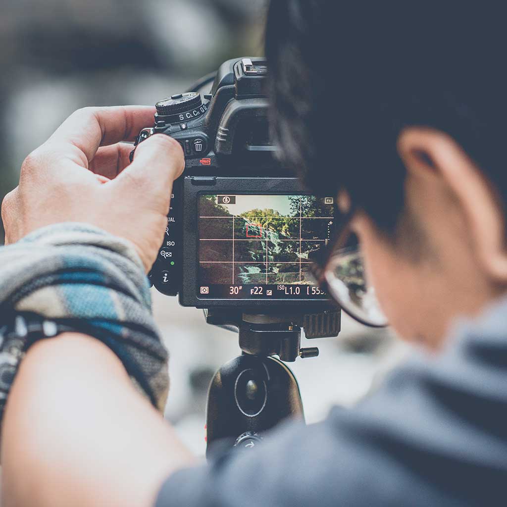 A photographer adjusting the settings on a camera while looking through the viewfinder. A tranquil rocky waterfall appears on the LCD screen.