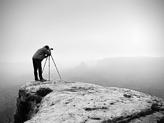 A photographer points a lense into the distance while standing on rock shelf. The colour scheme is black and white. The background is a mist covered mountainous landscape with a cloud covered sky.